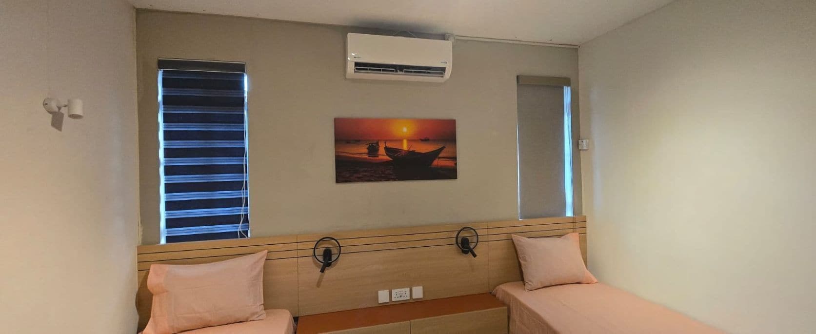 A room with two beds, an air conditioner, a painting of a sunset, and a window with blinds.