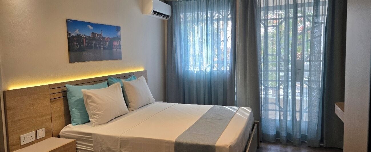 Habitación de hotel con cama, mesita de noche, aire acondicionado de pared, ventana con cortinas y una pintura de paisaje urbano.