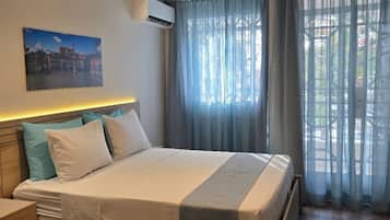 A hotel room with a bed, a nightstand, a wall-mounted air conditioner, a window with curtains, and a cityscape painting.