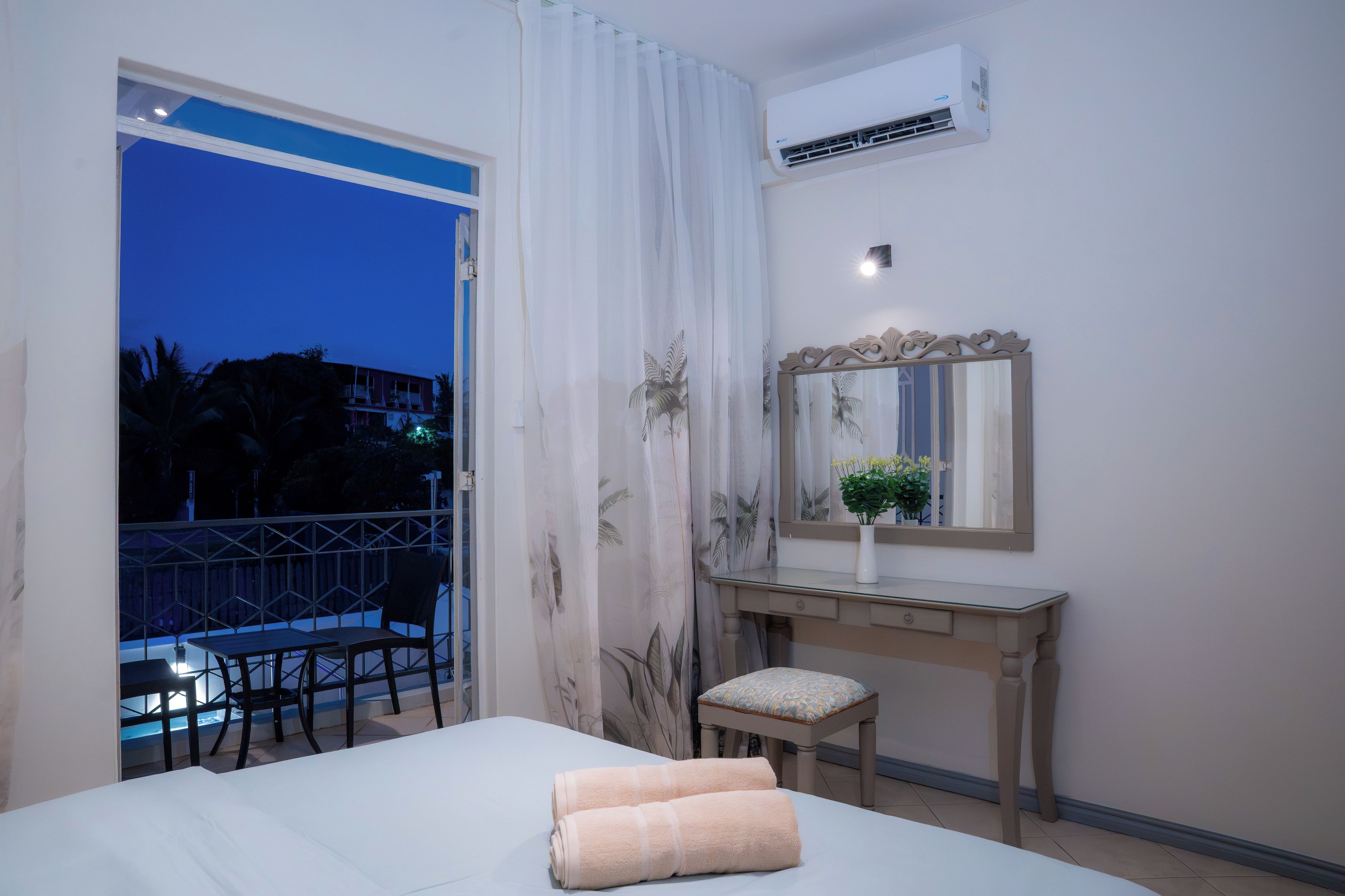 A hotel room with a bed, a desk, a chair, a mirror, and a balcony with a view of palm trees and a building.