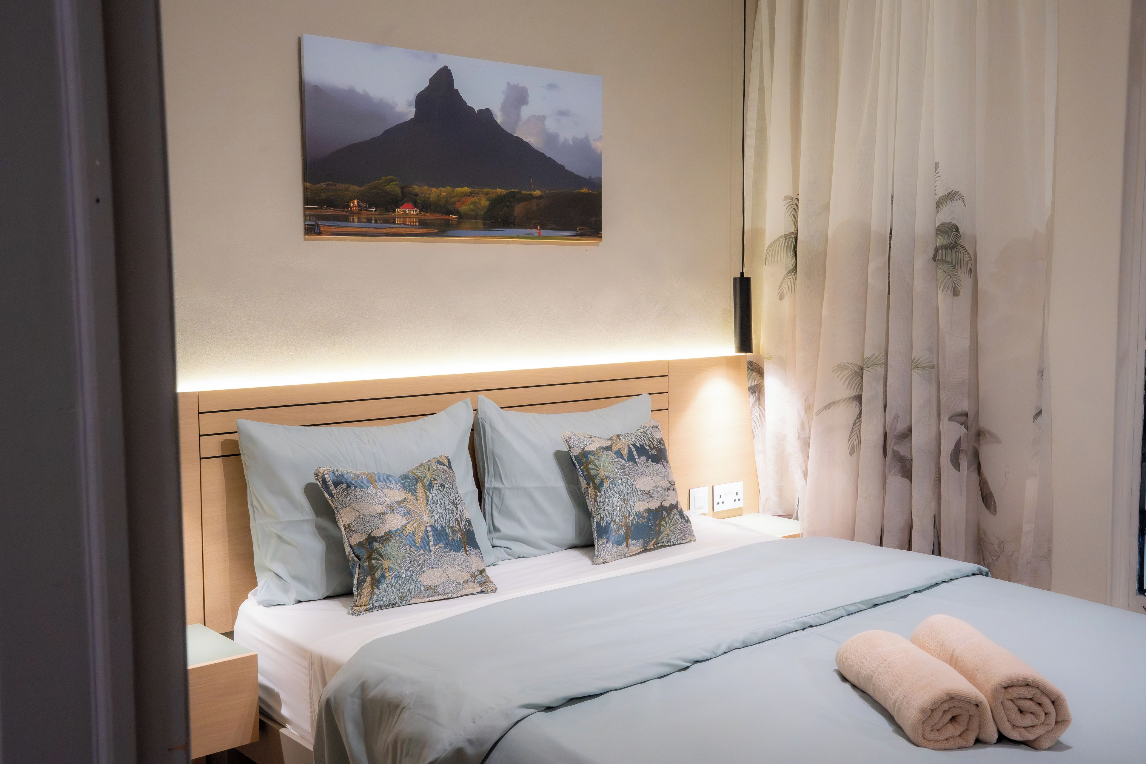 A neatly made bed with a light blue bedspread and pillows, a wooden headboard, and a painting of a mountain landscape above it.