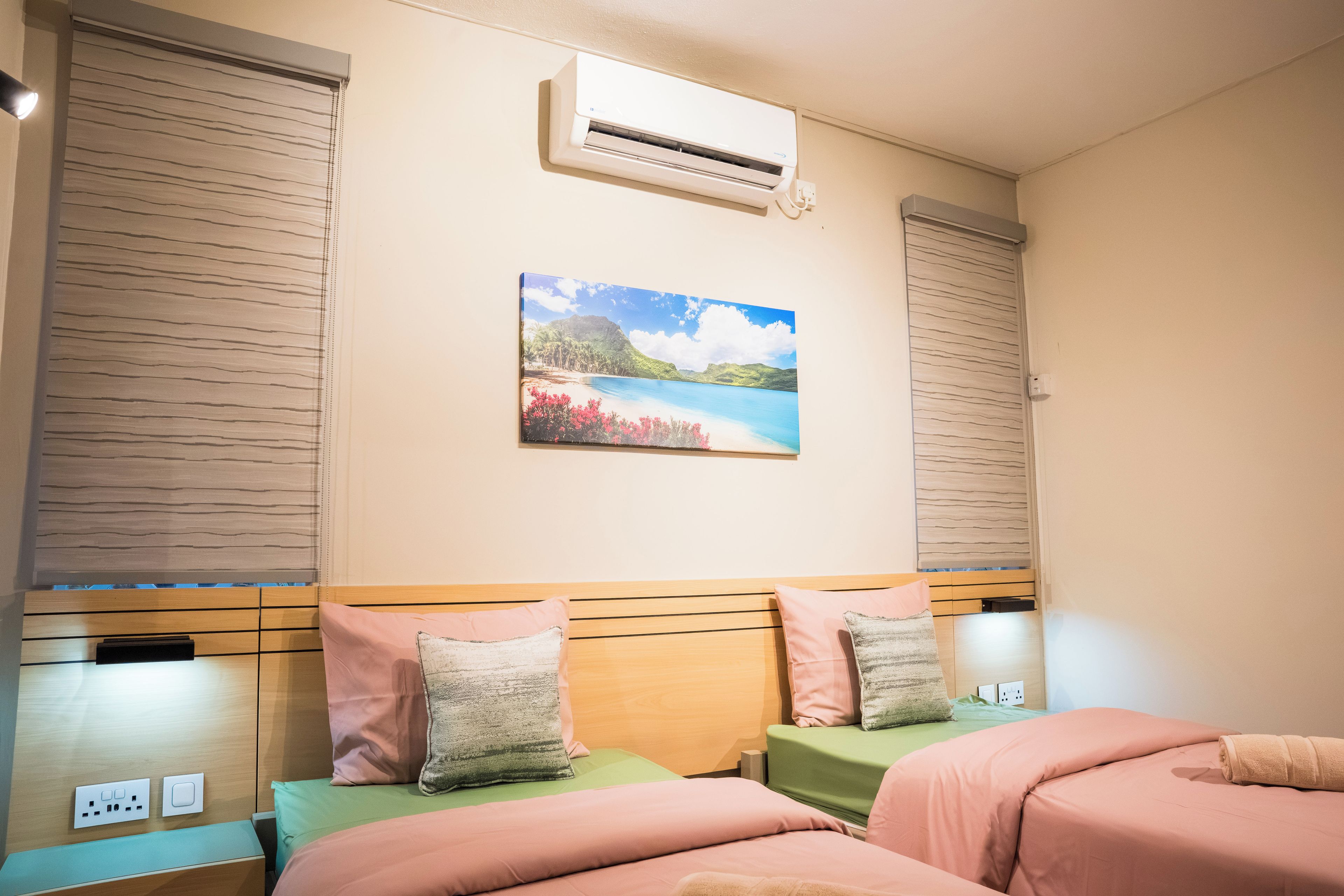A hotel room with a bed, a painting of a beach scene, an air conditioning unit, and a bedside table with a lamp.