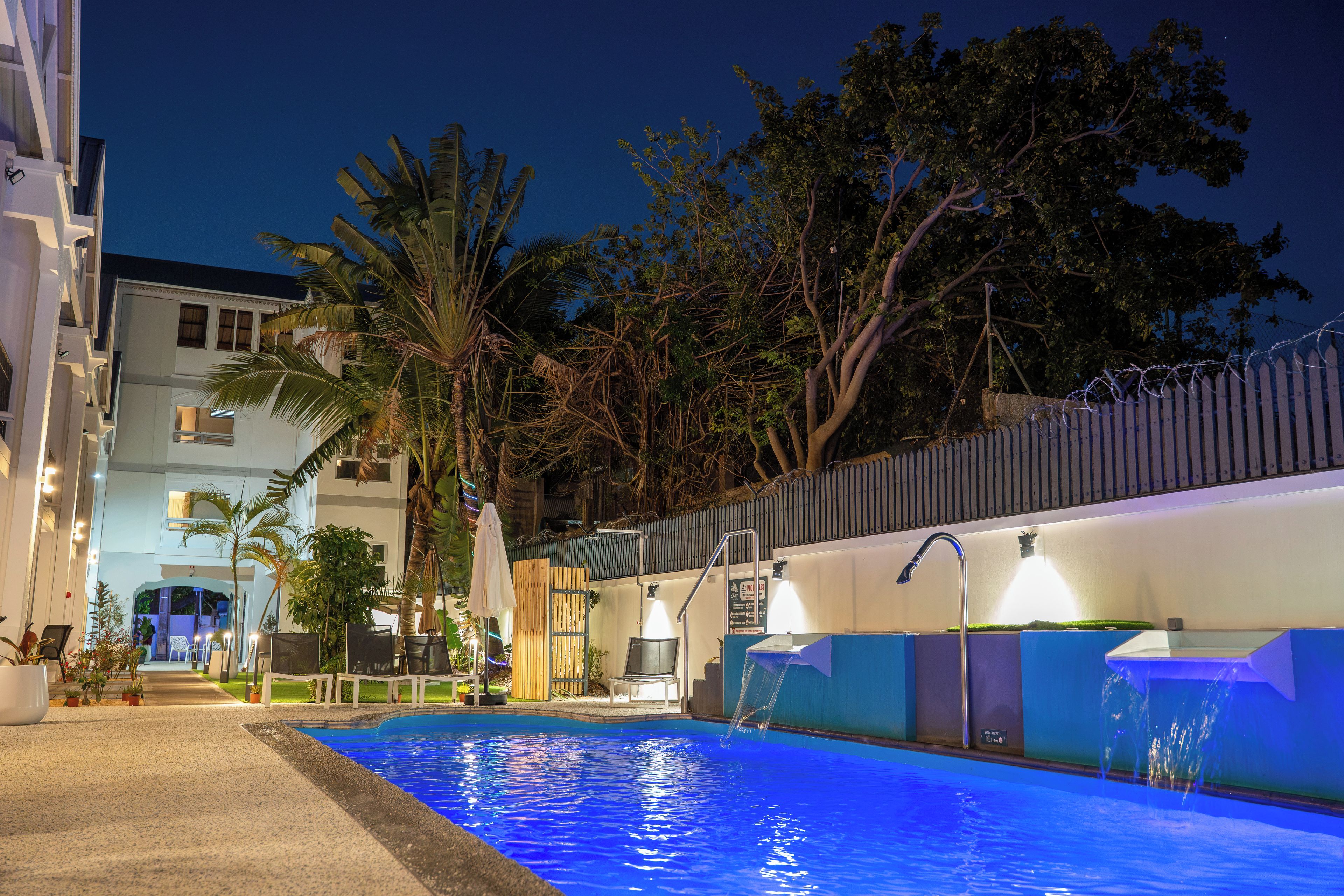 Ein Swimmingpool mit blauer Färbung, umgeben von einem Zaun, mit einem Gebäude und Bäumen im Hintergrund.