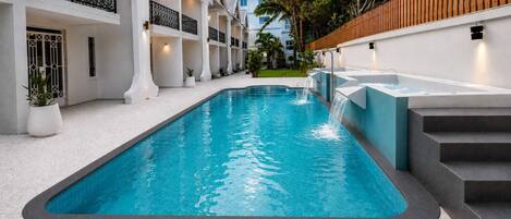 A swimming pool with a waterfall feature, surrounded by a white building with balconies and a wooden fence.