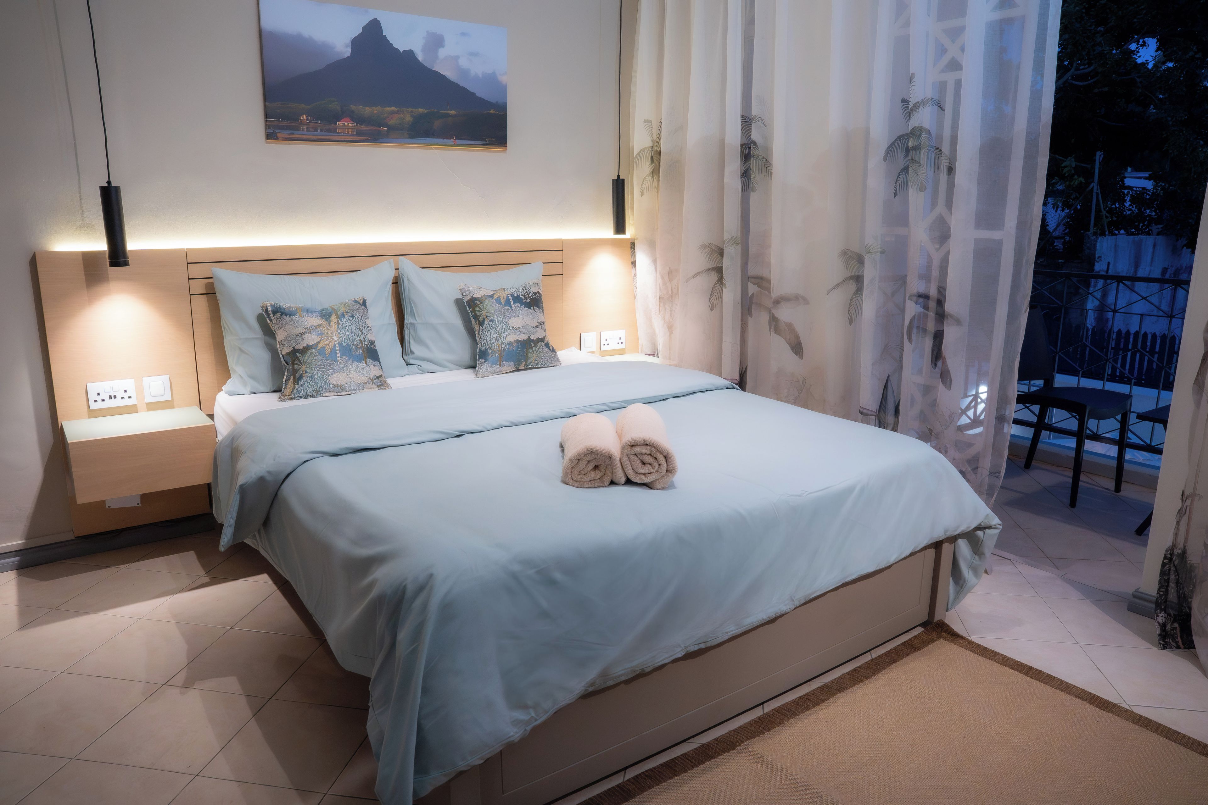 Una cama bien hecha con una colcha azul claro y almohadas, una mesita de noche con una lámpara y un cuadro de un paisaje montañoso sobre la cama.