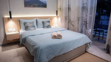 Una cama bien hecha con una colcha azul claro y almohadas, una mesita de noche con una lámpara y un cuadro de un paisaje montañoso sobre la cama.