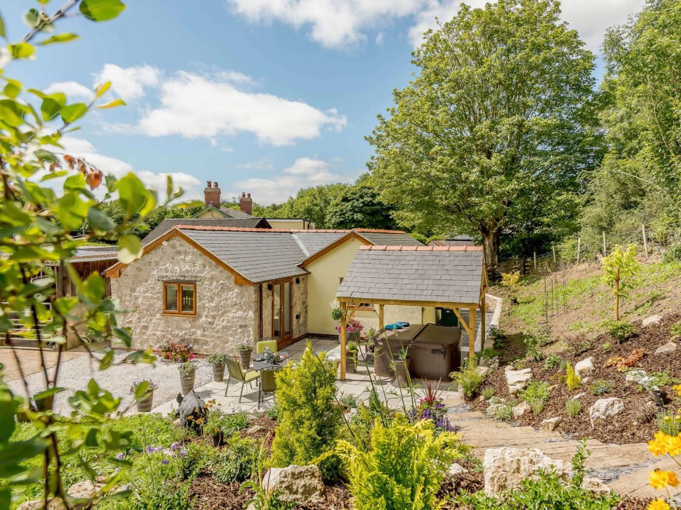 A stone house with a covered patio and a garden with various plants and a hot tub.
