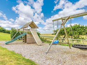 A playground with a wooden slide and swing set, surrounded by gravel and grass. - 3 Bed in St. Andrews (oc-88420) (Balmullo)