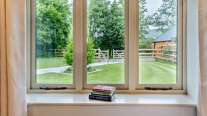 Ein Fenster mit drei Scheiben, das auf eine Wiese mit Holzzaun und ein Gebäude in der Ferne blickt.