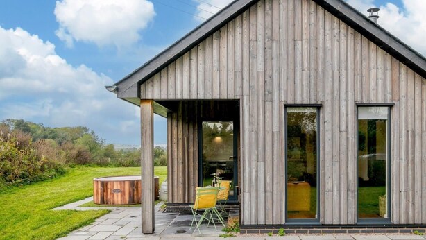 A modern wooden house with a covered patio, a hot tub, and a dining area.