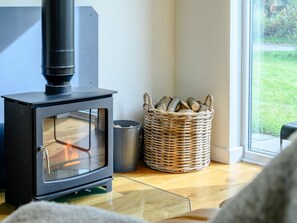 A room with a wood-burning stove, a wicker basket with logs, and a view of the outdoors through a window.