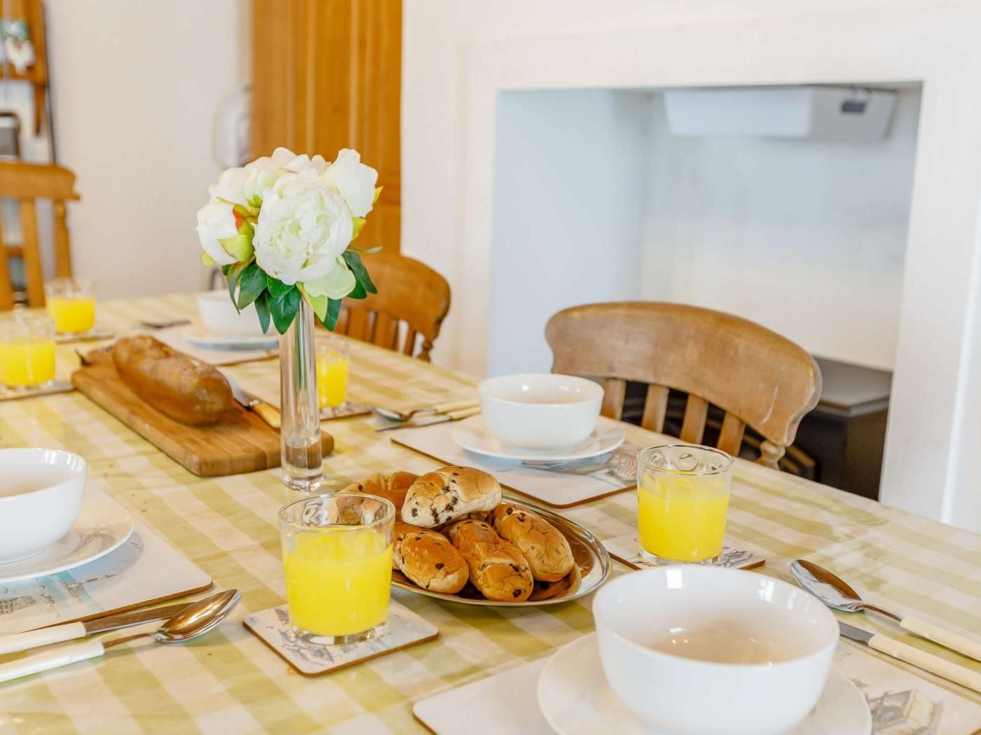 A breakfast table with a checkered tablecloth, a vase of flowers, a plate of pastries, glasses of orange juice, and a bowl of soup.
