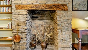 Una llar de pedra amb un mantell de fusta, una clau decorativa, una paperera de metall i un vas amb plantes seques.