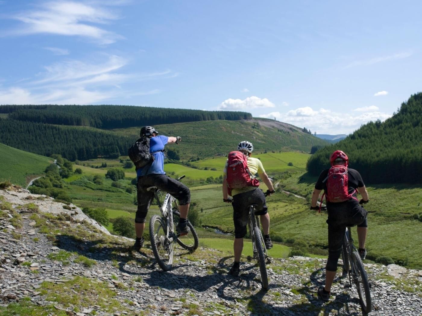 Trois cyclistes sur un sentier de montagne, avec vue sur une vallée et des collines boisées.