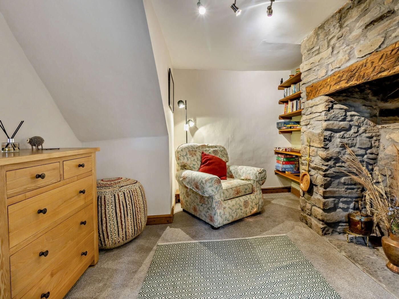 Un cuarto acogedor con un aparador de madera, un reposapiés estampado, un sillón con estampado floral, una chimenea de piedra y una estantería.