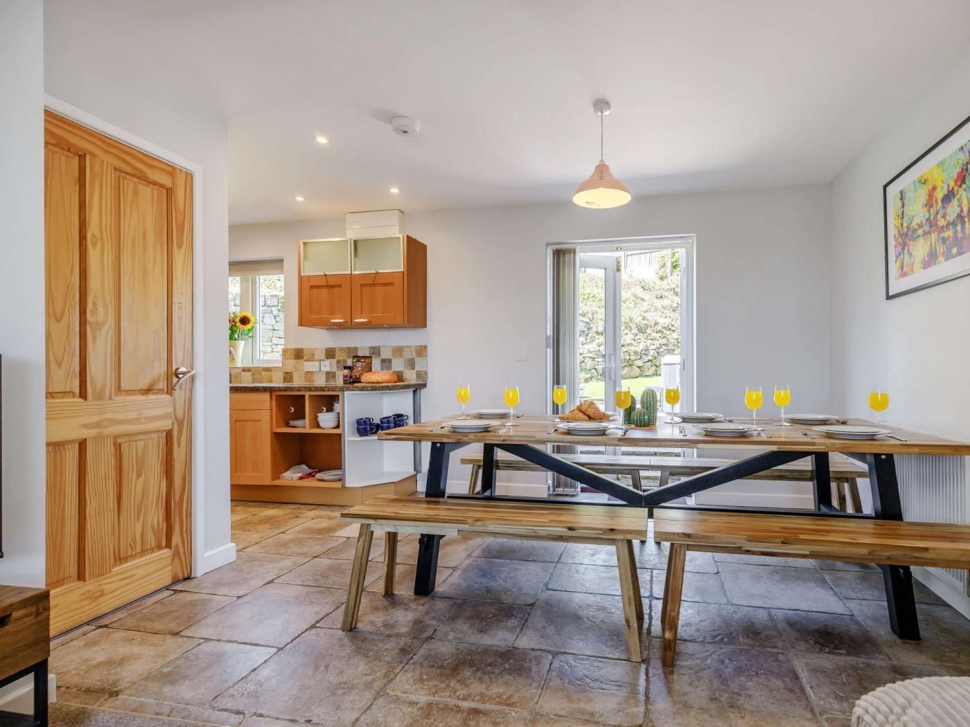 A modern kitchen with wooden cabinets, a stone-tiled floor, and a dining area with a bench and table set for four.