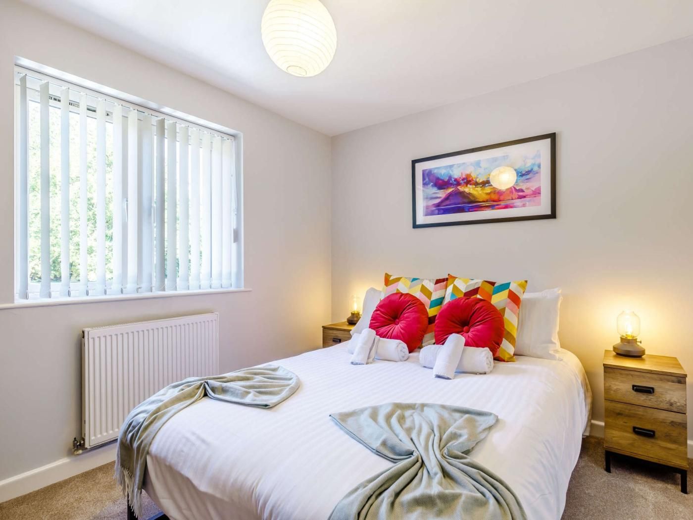 Une chambre à coucher avec un lit, un radiateur, une fenêtre avec des stores, une table de chevet avec une lampe et un tableau accroché au mur.