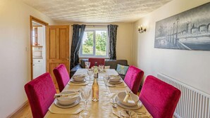Comedor con una mesa dispuesta para seis, una ventana con cortinas, un cuadro en la pared y una puerta de madera que da a la cocina.