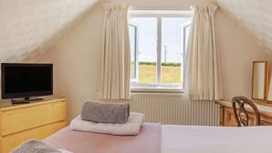 Un dormitorio con una televisión sobre un aparador de madera, una cama con una manta rosa, un ventanal con cortinas y vistas al exterior.