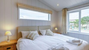 Ein ordentlich bezogenes Bett mit Kissen und einem gefalteten Handtuch, ein Nachttisch mit Lampe und ein Fenster mit Vorhängen.