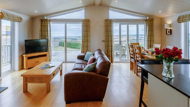 Ein geräumiger Wohnbereich mit einer Couch, einem Fernseher auf einem Holzboden, einem Esstisch mit Stühlen und Blick durch große Schiebetüren.