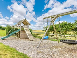 A playground with a wooden slide and swing set, surrounded by gravel and greenery. - 3 Bed in St. Andrews (oc-88545) (Balmullo)