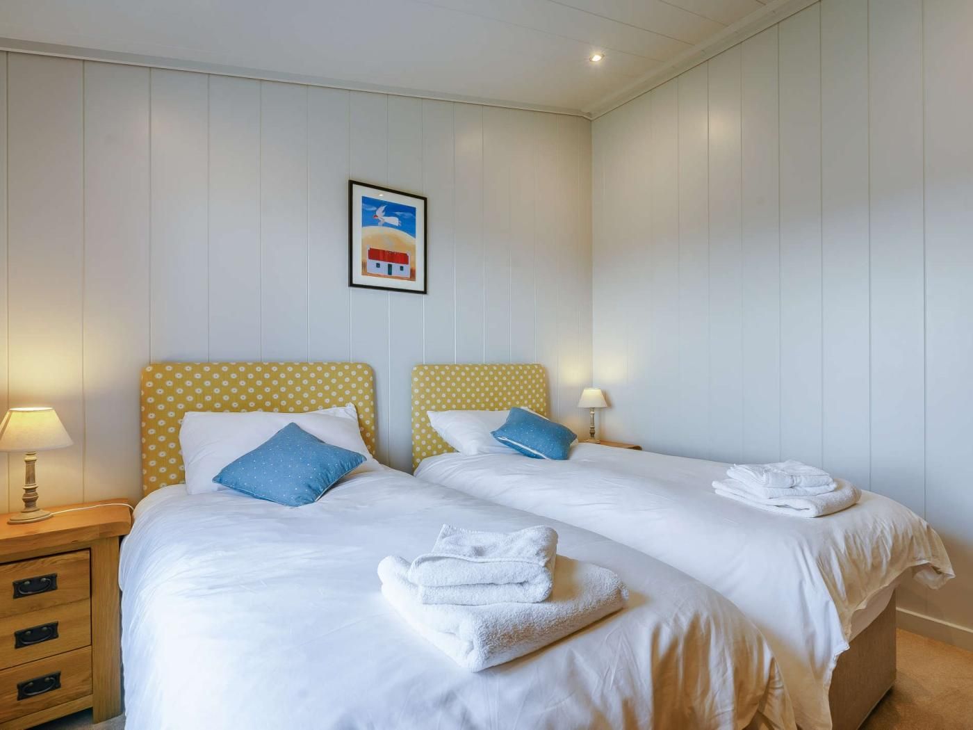 Une chambre d’hôtel avec deux lits, chacun doté d’une tête de lit jaune à pois, de draps blancs et d’un oreiller bleu. Un tableau encadré est accroché au mur, une table de chevet en bois avec une lampe s’y trouve, et un ensemble de serviettes est posé sur le sol.