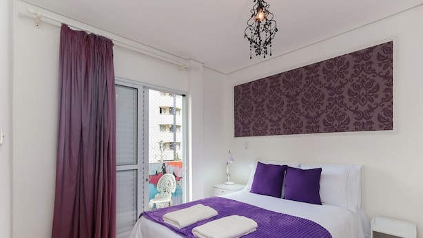 Un dormitorio con cama, ropa de cama morada, un candelabro, un papel tapiz estampado y una ventana con vistas.