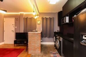 A compact living space with a brick fireplace, a washing machine, a refrigerator, and a television. - Apartamento p 4 com Varanda Prox Nubank Parque (São Paulo)