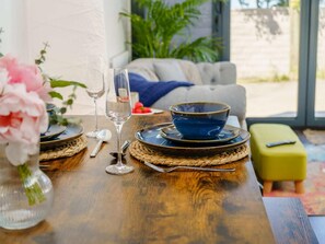 A dining table set with plates, bowls, cutlery, and glasses, with a vase of flowers and a green plant in the background.