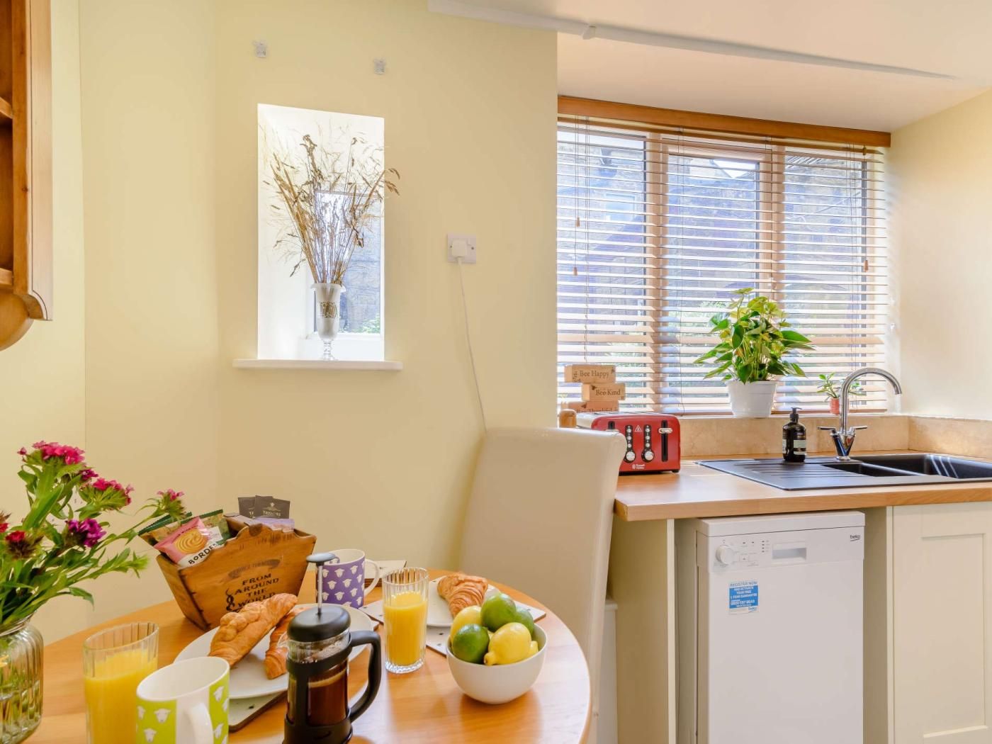 A kitchen with a sink, a window with blinds, a coffee maker, and a breakfast setup with croissants and juice.