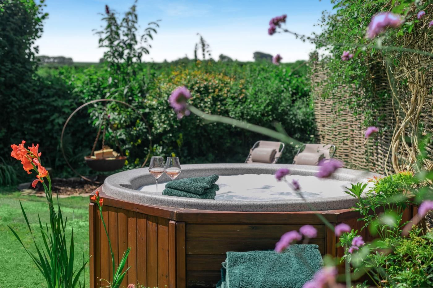 A hot tub with two glasses of wine, surrounded by greenery and a wooden deck.