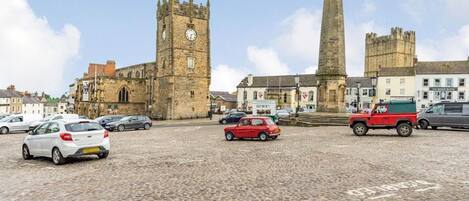 A cobblestone square with historic buildings, a clock tower, and a tall monument.