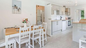 A modern kitchen with white cabinetry, a dining area with a wooden table and white chairs, and a view of the outdoors through a door. - 2 Bed in Saltash (oc-88313) (Notter)