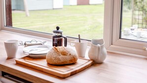 Mesa de café da manhã com cafeteira, xícaras, um prato e um biscoito em uma tábua de madeira, perto de uma janela com vista para um jardim e casas.