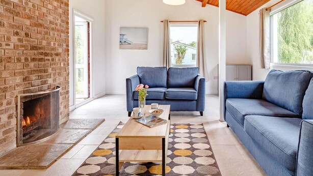 Uma sala de estar com lareira de tijolos, sofás azuis, uma mesa de centro de madeira e teto com vigas de madeira aparente.