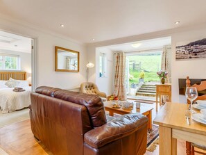 Ein geräumiges Zimmer mit Ledersofa, Esstisch, Stuhl, Bett, Spiegel und Blick auf einen Garten durch ein großes Fenster.