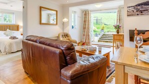 A spacious room with a leather sofa, a dining table, a chair, a bed, a mirror, and a view of a garden through a large window. - 1 Bed in Totnes (oc-88098) (North Huish)