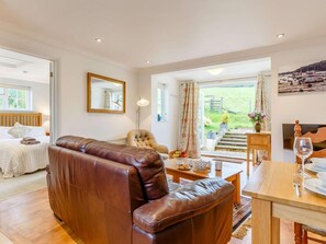 A spacious room with a leather sofa, a dining table, a chair, a bed, a mirror, and a view of a garden through a large window. - 1 Bed in Totnes (oc-88098) (North Huish)