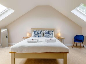 Ein Schlafzimmer mit schrägen Wänden, einem Bett mit weißer Bettwäsche, zwei Nachttischlampen, einem blauen Sessel und einem Spiegel.