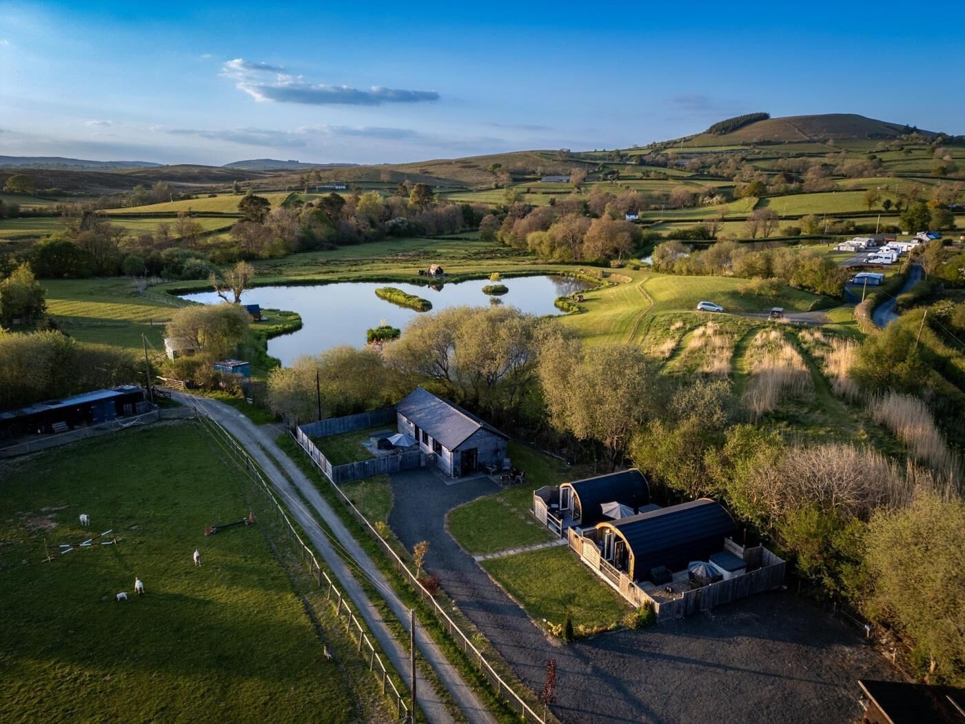 Un paisaje rural con un estanque, colinas onduladas y un edificio pequeño con un área cercada.