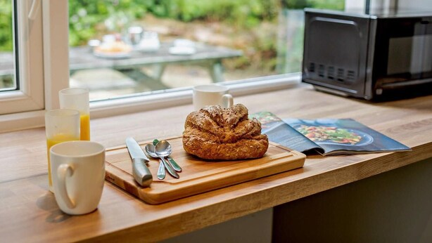 A breakfast setup with a croissant, two white mugs, a knife, and a spoon on a wooden table, with a window view of greenery and a microwave in the background. - 2 Bed in Saltash (oc-88310) (Notter)