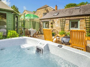 A hot tub in a garden with stone buildings, wooden chairs, and a table with a green umbrella. - 1 Bed in Bodmin (oc-88165) (Lanhydrock)