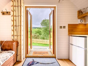 A room with a wooden door leading to a garden, a sofa, a refrigerator, and a wooden shelf with glassware.