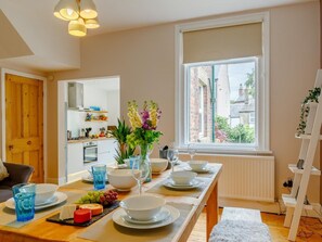 Un coin repas avec une table mise pour le repas, une fenêtre donnant sur des immeubles et une cuisine en arrière-plan.