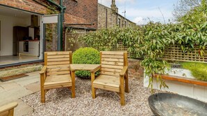 Une terrasse avec deux chaises en bois et une petite table, un sol en gravier, un bâtiment en briques et une haie.