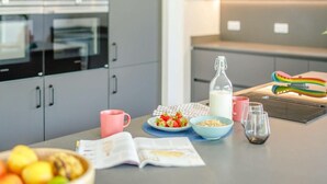 Une cuisine moderne avec des meubles gris, un four à micro-ondes, une bouilloire, une bouteille de lait, une tasse rose, un bol de fraises, un bol de céréales, un verre d’eau, un livre et un panier de fruits.