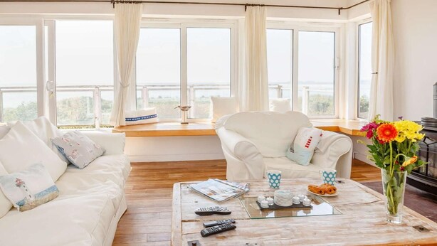Ein modernes Wohnzimmer mit einem weißen Sofa, einem weißen Sessel, einem Couchtisch mit Fernbedienungen und Blick ins Freie durch große Fenster.