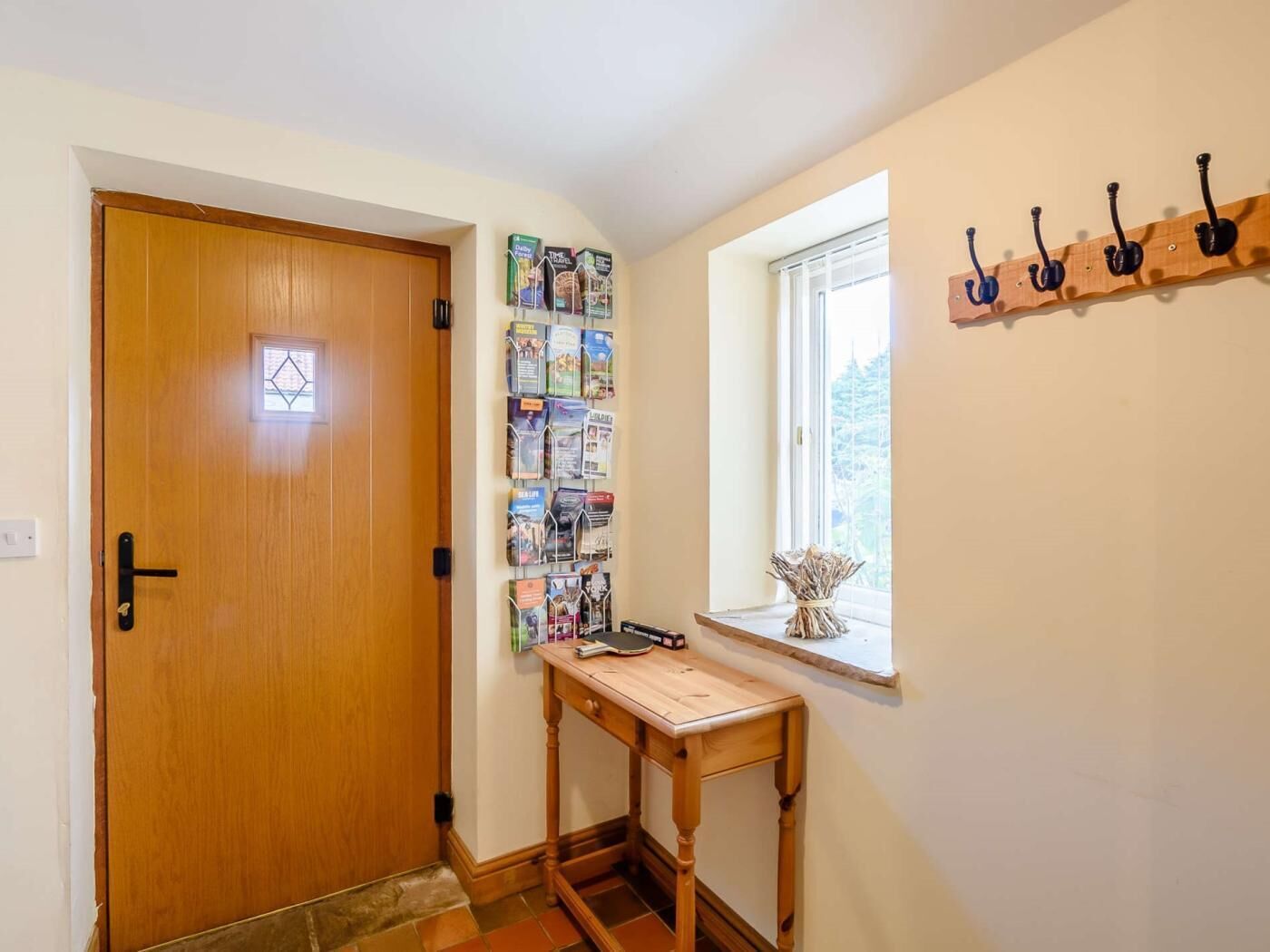 A hallway with a wooden door, a wall-mounted book rack, a small table, and a window with a view of trees.