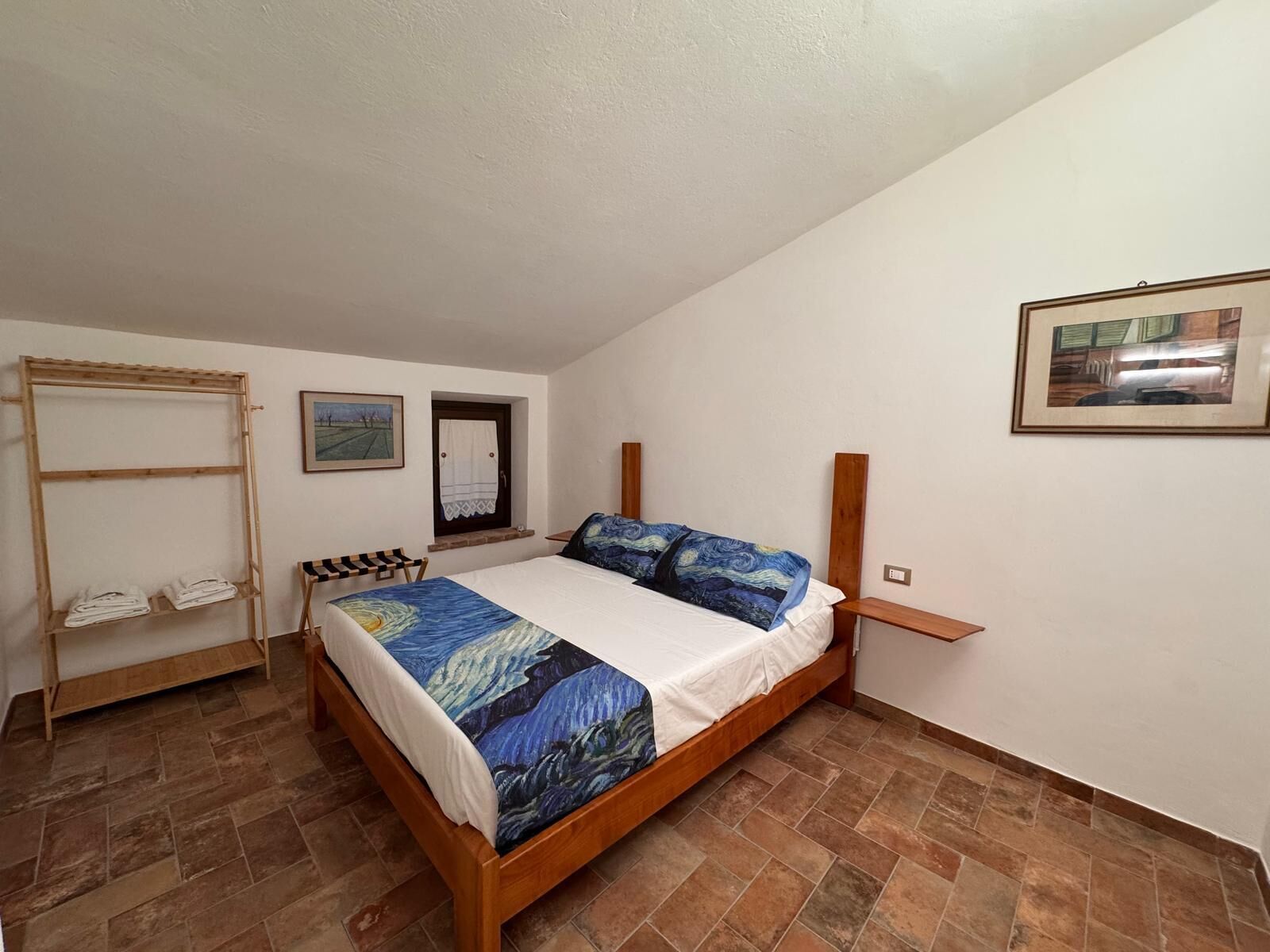 Un dormitorio con cama de madera, un tendedero de madera, una mesita y una ventana con cortina.
